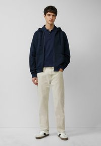 Granatowy hoodie na zamek z granatowym polo, w zestawieniu z kremowymi spodniami i białymi sneakersami z ciemnymi akcentami. Gładki materiał o luźnym kroju.