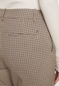 Close-up van beige en bruine houndstooth-gepatroонеerde broek met een achterzak en een klein rood, wit en blauw logo-label, gedragen met een crèmekleurige ribbeltrui.