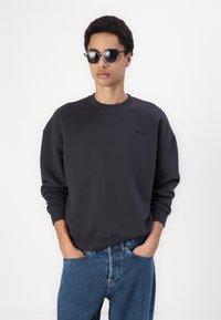 Mørkegrå hettegenser med rund hals, med en liten logo på brystet og avslappet passform. Kombinert med blå jeans og svarte solbriller.