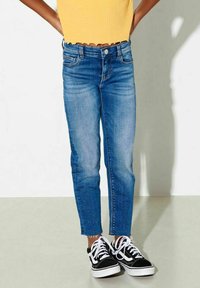 Jeans de denim azul con dobladillo deshilachado, diseño ajustado, cintura de subida media y un ligero desgaste, combinados con zapatillas blancas y negras.