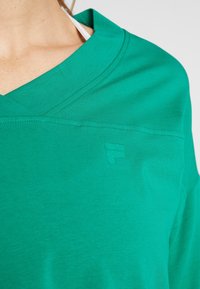 Zielona koszulka z dekoltem w serek, wykonana z miękkiej tkaniny; posiada subtelne logo wyhaftowane na klatce piersiowej oraz luźny krój. Gładka faktura bez wzorów.