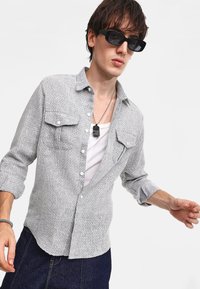 Camicia button-up in grigio a motivo houndstooth con due tasche sul petto, maniche lunghe, vestibilità rilassata e texture sottile. Indossata sopra una canotta bianca.