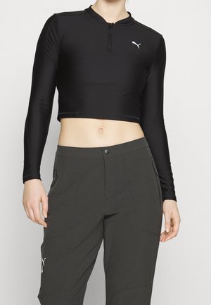 Kobieta ubrana w czarny długi rękaw krótki top z przednim zamkiem błyskawicznym oraz szare spodnie z wysokim stanem i kieszeniami na suwak po bokach.