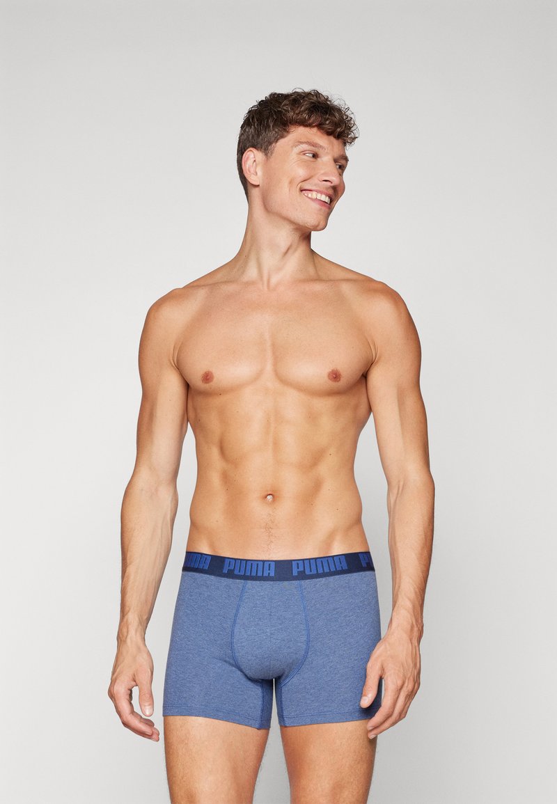 Man zonder shirt glimlacht en kijkt naar rechts, draagt blauwe Puma boxershorts met marineblauwe tailleband tegen een effen lichte achtergrond.