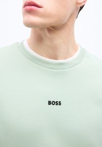 Νέος άνδρας που φοράει ανοιχτό πράσινο μπλουζάκι με στρογγυλή λαιμόκοψη και μαύρο κείμενο "BOSS" στο κέντρο του στήθους, κοιτώντας ελαφρώς προς το πλάι.