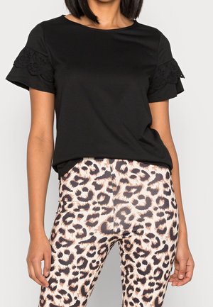 Vrouw draagt een zwarte top met kanten details aan de mouwen en leggings met luipaardprint, zichtbaar van midden torso tot midden dijen.