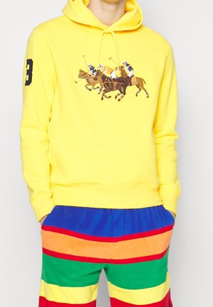 Homme portant un sweat à capuche jaune vif avec des joueurs de polo brodés et un short rayé coloré avec les mains dans les poches.