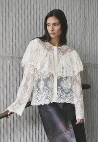 Lace blouse in ivory with ruffled sleeves and a sheer overlay. Features intricate floral patterns and a button closure at the front.