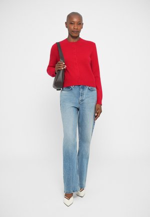 Pull rouge ajusté avec des boutons sur le devant ; associé à un jean flare bleu clair et des escarpins blancs à bout pointu ; sac à bandoulière noir.