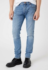 Homem a usar jeans slim-fit azul claro e botas pretas com atacadores, de pé contra um fundo branco liso.