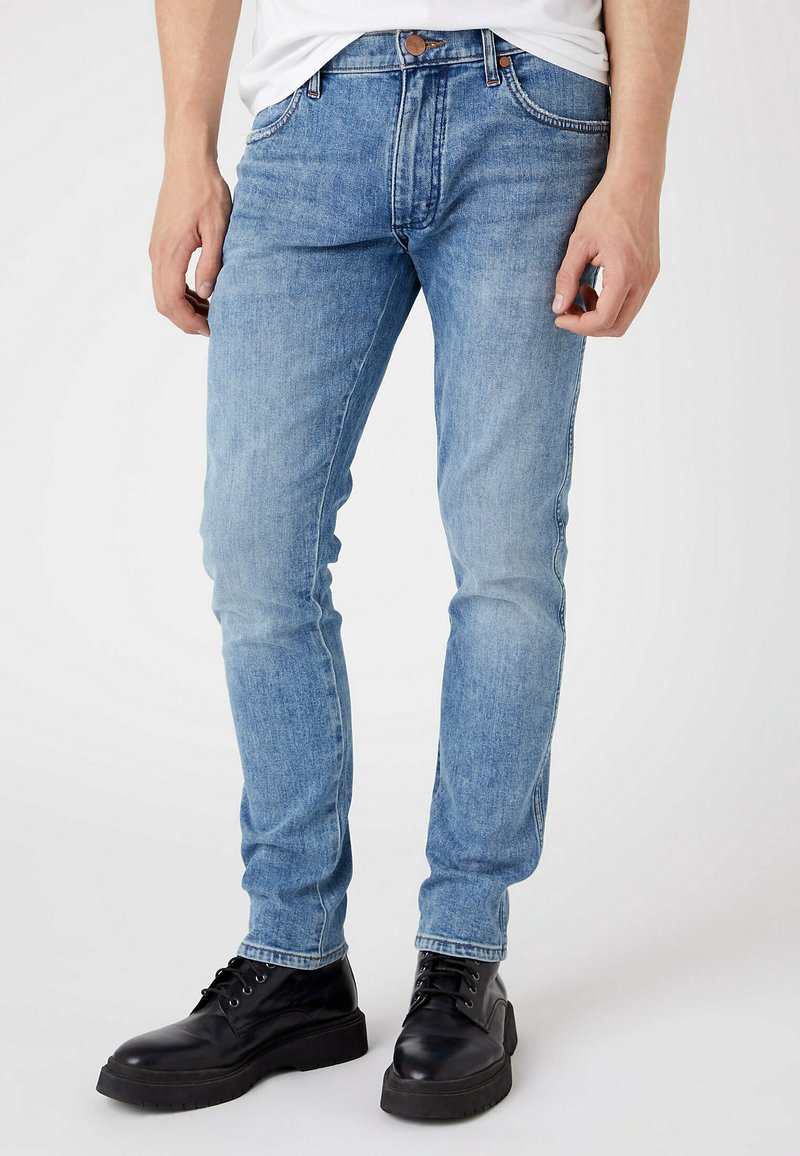 Homem a usar jeans slim-fit azul claro e botas pretas com atacadores, de pé contra um fundo branco liso.