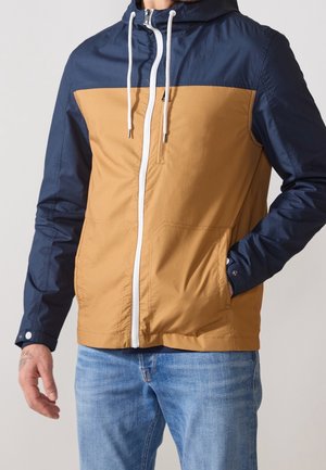 Hombre con una chaqueta con capucha bicolor con mangas y parte superior azul marino, cuerpo color canela, cremallera blanca y vaqueros azules, de pie con una mano en el bolsillo.