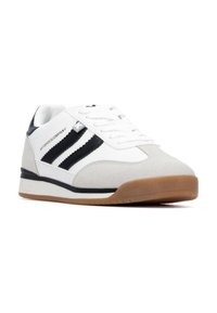 Zapatilla con una parte superior de tela blanca, acentos de gamuza gris, rayas negras y una suela de goma color caramelo. Presenta cordones blancos y un logo en el lateral.