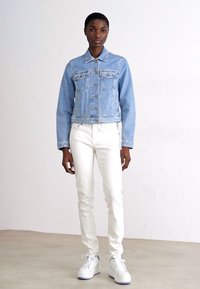 Giacca di jeans azzurro chiaro, aderente, con tasche sul petto, indossata sopra jeans bianchi slim-fit e sneakers bianche. Texture liscia, design semplice.