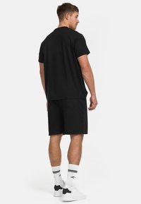Mand står med ryggen til iført en sort t-shirt, sorte shorts, hvide ankelstrømper med sorte striber og hvide sneakers.