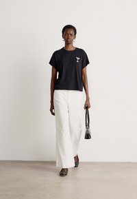 Sort bomulds T-shirt med et lille cocktailglas grafik, parret med hvide vide bukser og en sort håndtaske. Modellen har sorte sko på.