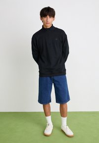 Sudadera con cremallera negra de tela de forro polar, combinada con pantalones cortos de mezclilla. El modelo lleva calcetines blancos y zapatos color tostado, de pie sobre un suelo verde.