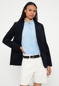 Morski sztruksowy blezer na jasnoniebieskim rozpinanym swetrze, w połączeniu z białymi szortami i czarnym paskiem. Gładkie tekstury i dopasowany krój.