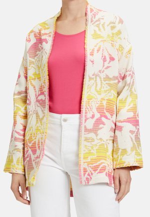 Femme portant un pantalon blanc, un top rose sans manches et un cardigan à fleurs multicolore ouvert aux tons roses et jaunes.