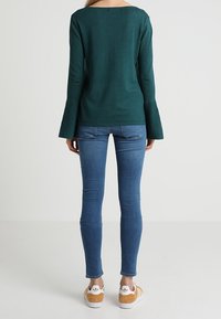 Zelený top s dlouhým rukávem s rozšířenými manžetami, v kombinaci s modrými úzkými džíny a béžovými teniskami s bílými doplňky. Zobrazen pohled zezadu.