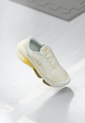 Scarpa da corsa Nike di colore chiaro con dettagli gialli e unità ammortizzanti visibili, mostrata a mezz'aria su uno sfondo chiaro e strutturato.