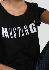 Osoba w czarnej koszulce z wyblakłym białym logo "MUSTANG" i ciemnoniebieskich dżinsach, skróconych u ramion i w talii.