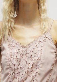 Camisole rose pâle avec un panneau en dentelle à motifs floraux, bordure délicate en feston le long de l'encolure et des bretelles, fabriquée en tissu léger.