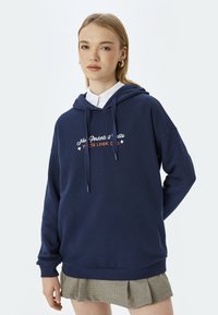 Niebieska bluza z kapturem z miękkiego materiału, z sznurkiem. Napisy z przodu w białym i pomarańczowym kolorze. Połączona z krótką, plisowaną spódnicą w kratę.