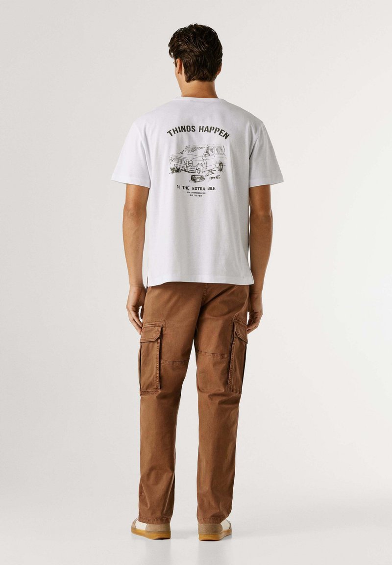 Wit katoenen t-shirt met "THINGS HAPPEN" afbeelding op de rug, gecombineerd met bruine cargobroek met zakken aan de zijkanten.