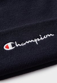 Beanie lavorato a maglia blu navy con una texture morbida. Presenta il logo "Champion" ricamato in bianco, accentuato da un'emblema rosso e bianco.