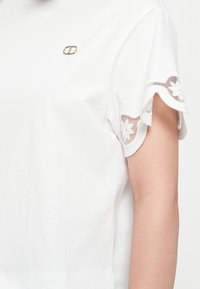 White cotton t-shirt with floral lace trim on sleeves and a small gold logo emblem on the chest. Short sleeves, casual fit.