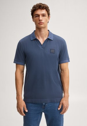Young man standing wearing a blue short-sleeve polo shirt and blue jeans against a plain light background.