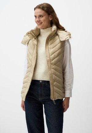 Beige gesteppte Weste mit hohem Kragen und Druckknöpfen, ausgestattet mit einem Reißverschluss und seitlichen Taschen, getragen über einem cremefarbenen Pullover und dunklem Denim.