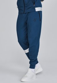 Navy Sweatpants mit elastischen Bündchen, die ein weißes Akzentband am Knöchel und ein aufgenähtes Logo am Oberschenkel aufweisen. Das Material wirkt weich.