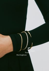 Ruka s tri narukvice: tanka zlatna lančana narukvica ribljeg kostura, tenis narukvica s dijamantima i zlatni polumjesečni narukvica na crnom rukavu.