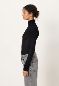 Kihara juustega naine kuklas, seljas must kõrge kaelusega kampsun ja hallid kõrge vöökohaga teksapüksid, seisab külje pealt valge tausta ees.