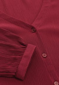 Blusa rossa con texture, dotata di scollo a V, chiusure a bottoni e maniche plissettate. Il materiale sembra leggero e leggermente stropicciato.