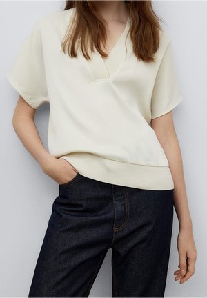 Kobieta w kremowej bluzce z krótkim rękawem i dekoltem w serek oraz ciemnoniebieskich wysokich jeansach, z jedną ręką w kieszeni.