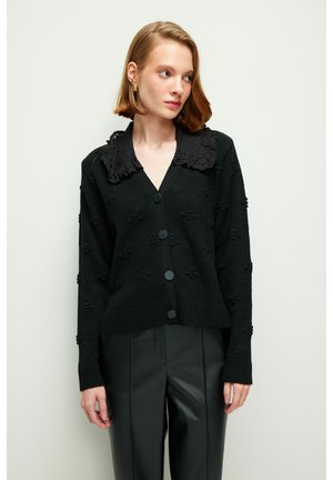 Schwarze Strickjacke mit gewelltem schwarzen Kragen, Knopfverschluss und strukturiertem Muster. Getragen über schwarzen Lederhosen.