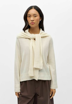 Femme aux longs cheveux foncés porte un pull crème avec une écharpe assortie drapée sur les épaules et un pantalon ample marron foncé sur fond blanc.