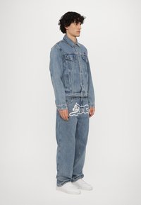 Giacca di denim con molte tasche, lavaggio blu e chiusura con bottoni. Abbinata a jeans a vestibilità ampia con un design grafico bianco.