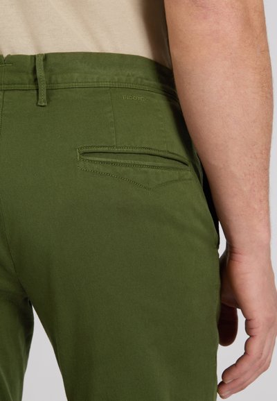 Close-up van een persoon die olijfgroene broek draagt met een achterzak met bies en een beige overhemd ingestopt, hand ontspannen langs het lichaam.