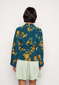 Blommig långärmad blus i teal med orange blommor och gröna stjälkar, med en rynkad halsringning och en cinchad midja. Åtföljd av en ljusgrön kjol.