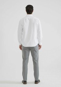 Chemise blanche à manches longues avec poignets à boutons et ourlet incurvé, associée à un pantalon gris clair, vue de dos.