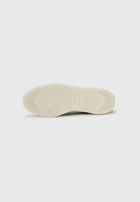 Sålen av en sneaker vist ovenfra, med et teksturert design med spor og en lys kremfarge på en ren hvit bakgrunn.