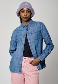 Camisa de denim en azul claro con botones en la parte delantera, dos bolsillos en el pecho y botones metálicos, llevada sobre una camiseta negra y combinada con pantalones rosas.