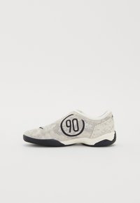 Zapatilla de malla gris con acentos negros y un logotipo circular que muestra "90". Presenta un diseño de perfil bajo y un sutil patrón texturizado.
