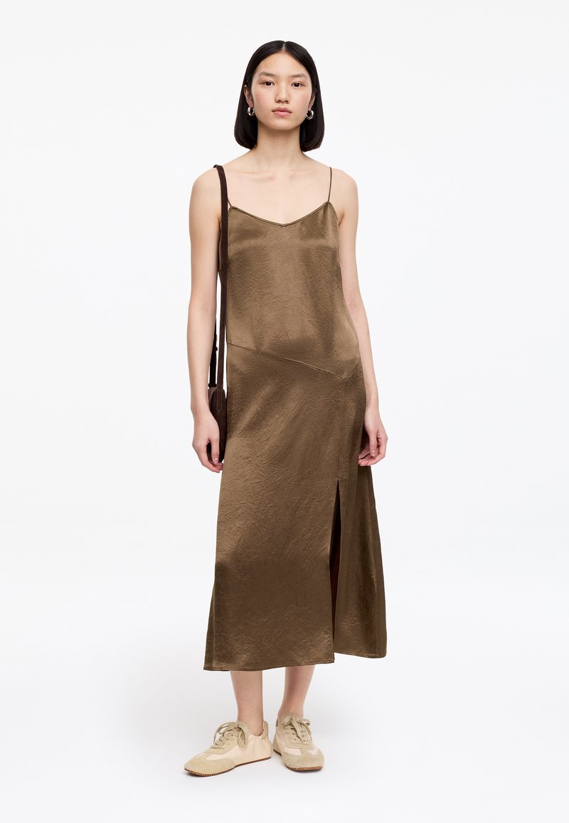 Jeune femme portant une robe nuisette en satin marron avec une fente latérale, des baskets beiges, des boucles d'oreilles créoles, et un petit sac à bandoulière marron.