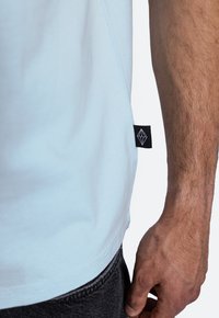 Světle modré tričko s krátkým rukávem a hladkým povrchem. Na bočním švu se nachází černá etiketa s bílým logem a v blízkosti jsou viditelné šedé džíny.