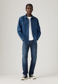 Camicia in denim blu scuro con bottoni, dotata di due tasche sul petto, indossata sopra una T-shirt bianca. Abbinata a jeans azzurri chiari e sneaker nere.